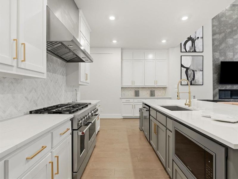 Contemporary kitchen featuring white cabinetry with gold-tone hardware, a stainless steel range with matching hood, a white herringbone tile backsplash, and a large central island with a built-in microwave and sink Contemporary kitchen featuring white cabinetry with gold-tone hardware, a stainless steel range with matching hood, a white herringbone tile backsplash, and a large central island with a built-in microwave and sink