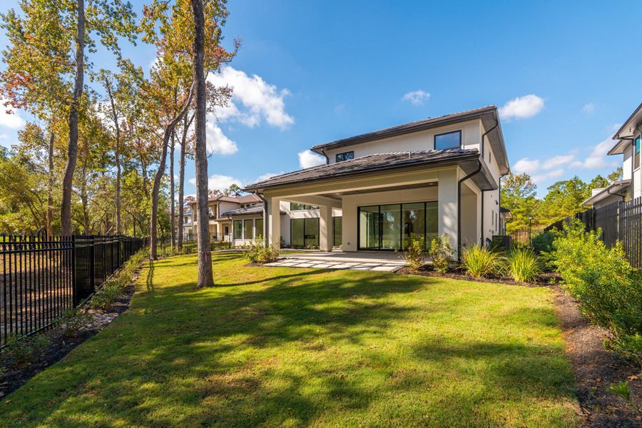 This photo showcases a modern two-story home with a spacious, well-maintained backyard. The house features large windows and a covered patio, perfect for outdoor entertaining. The yard is bordered by a sleek black fence and surrounded by mature trees, providing a serene setting.