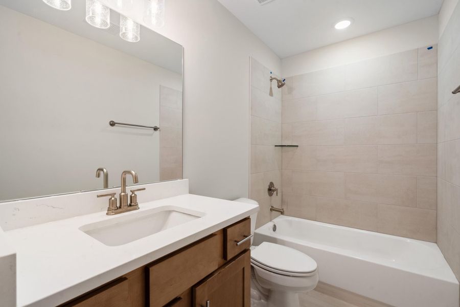 Bathroom featuring a white bathtub with light-colored tile surround, a single vanity with wood cabinetry, a white countertop, and a rectangular undermount sink Bathroom featuring a white bathtub with light-colored tile surround, a single vanity with wood cabinetry, a white countertop, and a rectangular undermount sink