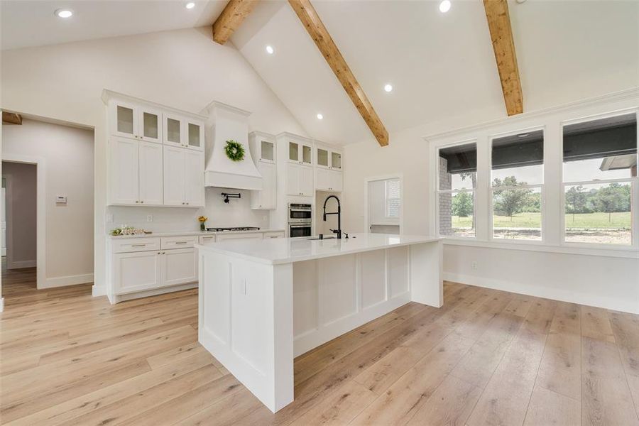 Bright kitchen featuring a large central island with a matte black faucet, white shaker cabinetry, wood-finish flooring, a vaulted ceiling with exposed wood beams, and a wall of windows