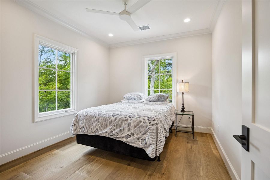 Bedroom of the light-filled mother-in-law/guest suite featuring French white oak wide plank flooring, crown molding, recessed lighting and ceiling fan, along with large windows that bring in abundant natural light and treetop views. A clean, airy palette creates a welcoming and comfortable retreat.