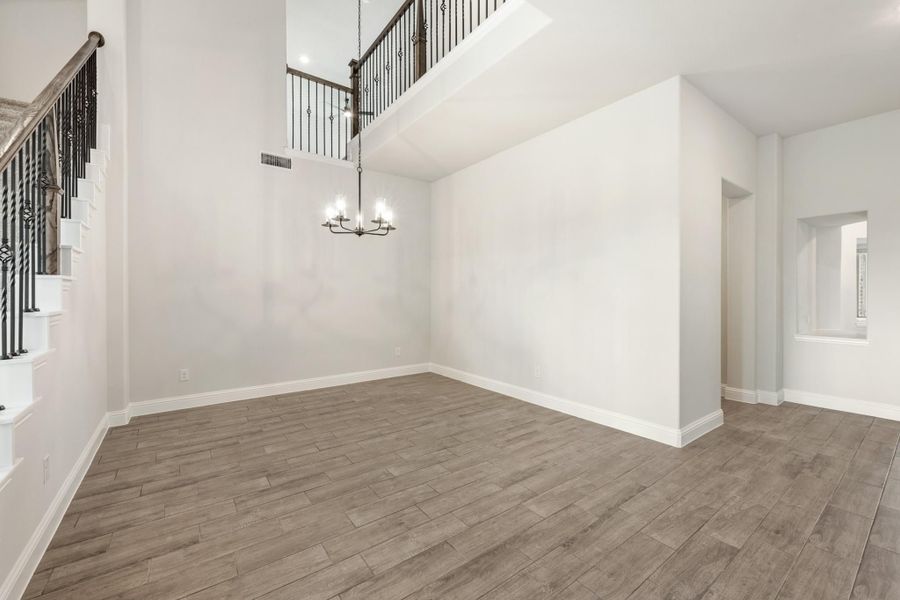 Empty living room with two-story ceiling, chandelier, wood-look floors, and wrought iron staircase railing above. Empty living room with two-story ceiling, chandelier, wood-look floors, and wrought iron staircase railing above.