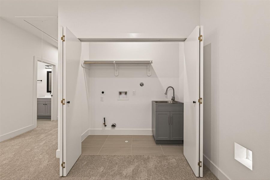 This photo shows a laundry area with open double doors revealing hookups for a washer and dryer, a small sink with a cabinet, and a shelf for storage. The room has carpeted flooring leading to a tiled section. This photo shows a laundry area with open double doors revealing hookups for a washer and dryer, a small sink with a cabinet, and a shelf for storage. The room has carpeted flooring leading to a tiled section.