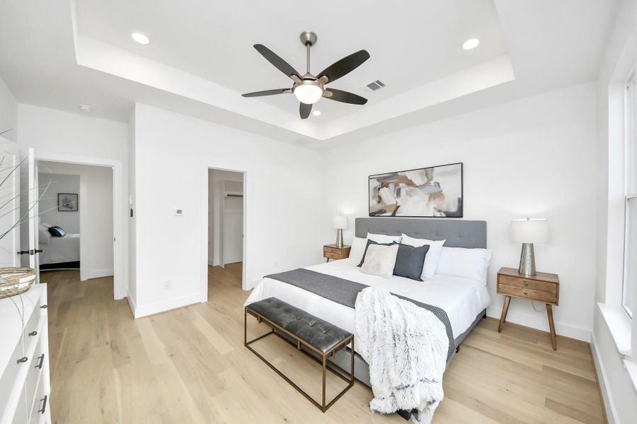 This spacious bedroom features a modern design with a tray ceiling and recessed lighting. It includes a large bed with stylish bedding, complemented by two wooden nightstands and lamps. The room is bright with natural light, thanks to large windows, and has light hardwood flooring. A ceiling fan adds to the comfort, and there's a walk-in closet for ample storage.