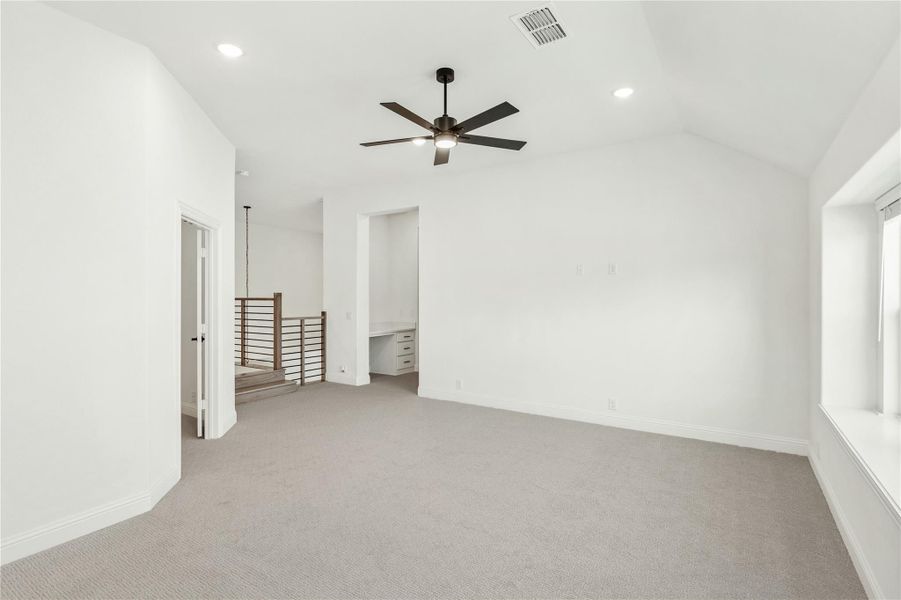 Empty bedroom with vaulted ceiling, ceiling fan, carpet flooring, and built-in desk area near staircase railing