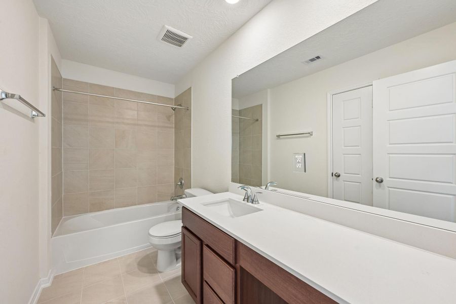 The secondary bathroom offers tile flooring, a bath/shower combination with a stylish tile surround, rich stained wood cabinetry with added knee space and elegant light countertops, a mirror, and sleek fixtures with modern finishes.