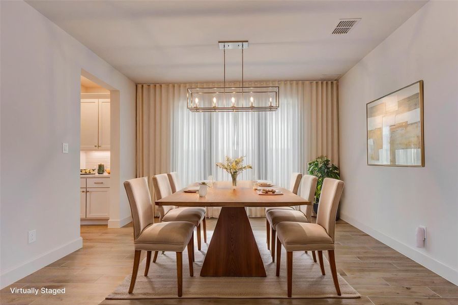 Dining room with a modern chandelier The room features light wood looking tile flooring, neutral walls, and a large window with sheer curtains for natural light. Adjacent to the dining area is a glimpse of a contemporary kitchen. Dining room with a modern chandelier The room features light wood looking tile flooring, neutral walls, and a large window with sheer curtains for natural light. Adjacent to the dining area is a glimpse of a contemporary kitchen.