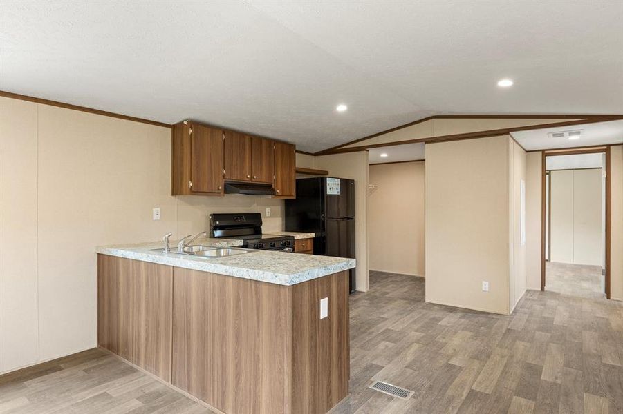 Kitchen area featuring wood-finish flooring, a kitchen island with a double basin sink and light-toned countertop, wood cabinetry, and black appliances including a range and refrigerator