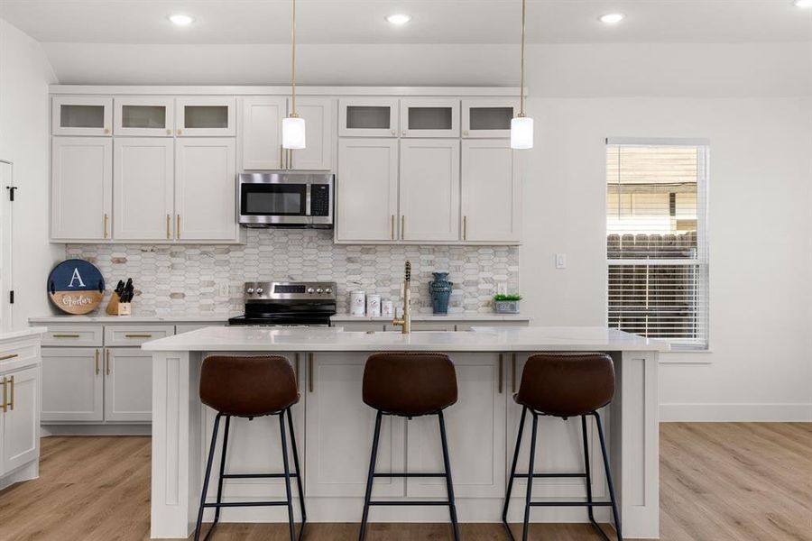 Kitchen with light hardwood / wood-style flooring, appliances with stainless steel finishes, hanging light fixtures, an island with sink, and white cabinets