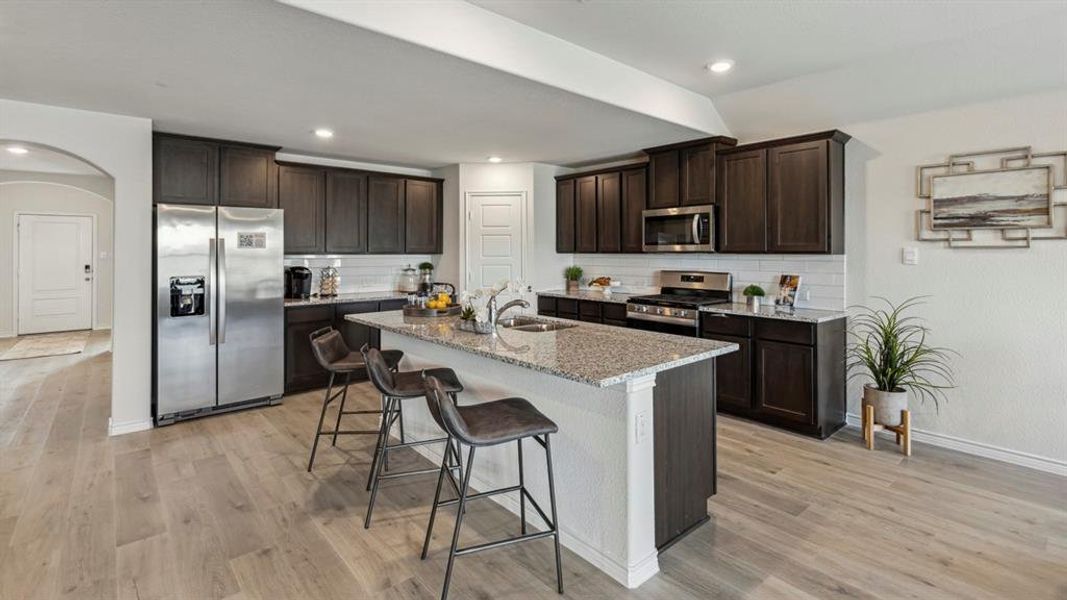 The kitchen features dark wood cabinetry, stainless steel appliances, a large island with a light-colored granite countertop, and light wood-look flooring