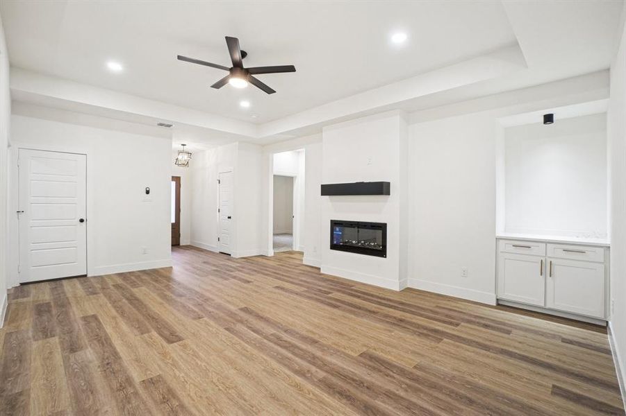 Unfurnished living room with light wood finished floors, a tray ceiling, a glass covered fireplace, recessed lighting, and ceiling fan Unfurnished living room with light wood finished floors, a tray ceiling, a glass covered fireplace, recessed lighting, and ceiling fan