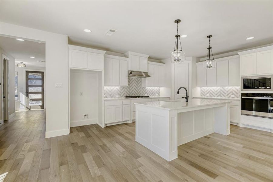 Kitchen featuring stainless steel oven, decorative light fixtures, white cabinetry, and an island with sink