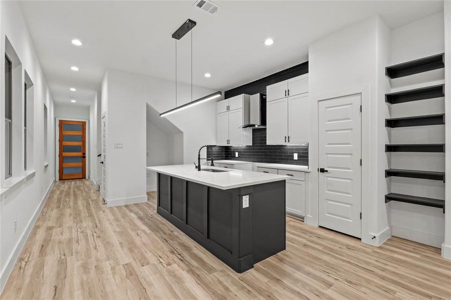 Kitchen featuring white cabinetry, wall chimney range hood, pendant lighting, light hardwood / wood-style floors, and a center island with sink