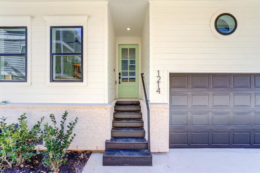 This photo showcases a modern home entrance with a stylish light green door, black accented double paned windows, adjacent to a matching garage door. The exterior features clean white Hardie Plank siding and a small landscaped area with shrubs.