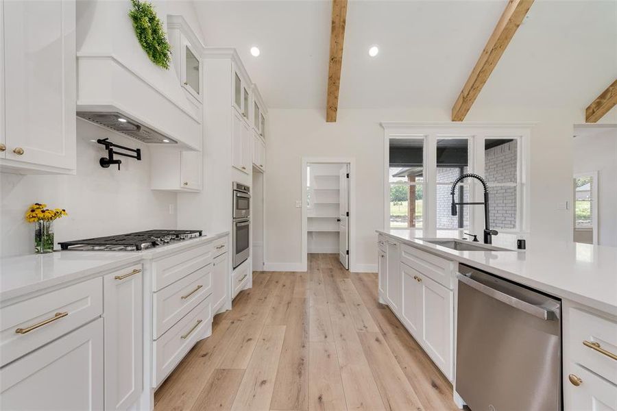 Modern kitchen featuring white cabinetry, light wood-finish flooring, exposed ceiling beams, and integrated stainless steel appliances
