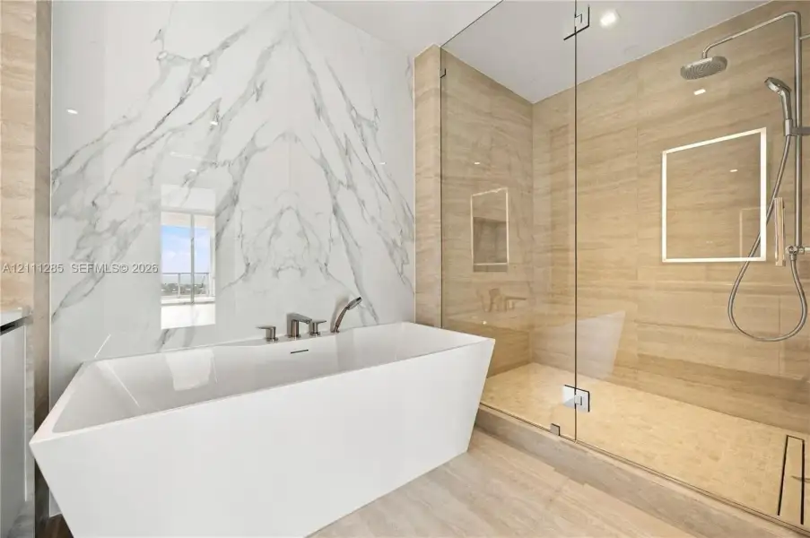 Freestanding soaking tub with a wall-mounted faucet, large-format stone-tile wall, glass-enclosed shower, integrated rainfall showerhead, and recessed lighting Freestanding soaking tub with a wall-mounted faucet, large-format stone-tile wall, glass-enclosed shower, integrated rainfall showerhead, and recessed lighting
