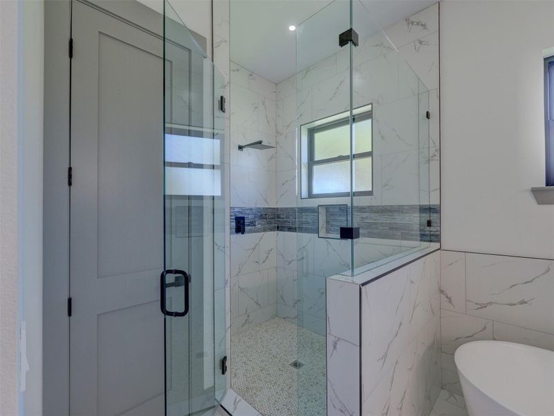Custom tile work accents a modern glass-enclosed shower featuring elegant marble patterns and neutral finishes.