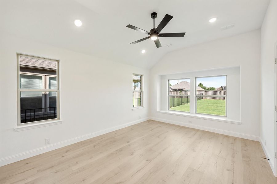 Empty bedroom with vaulted ceiling, black ceiling fan, light hardwood floors, and multiple windows with backyard views.