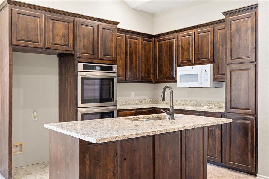 Kitchen featuring a center island with sink, dark wood finish cabinets, stainless steel double oven, white microwave, and light stone counters