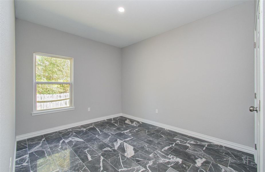 This room features a neutral color palette with gray walls and striking black marble-like flooring. A single window lets in natural light, and there is a simple light fixture on the ceiling. The space is minimalistic and versatile, suitable for various uses.