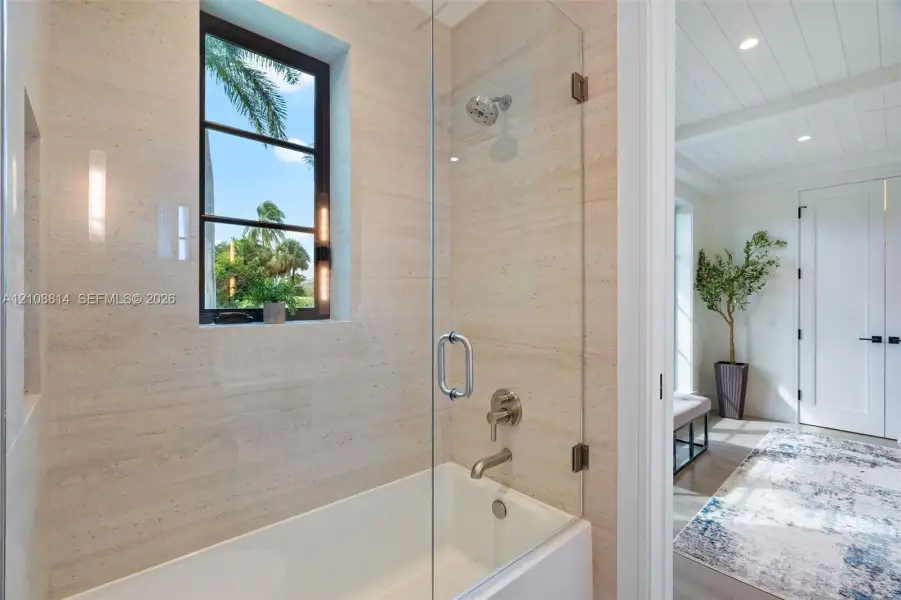 Bathroom featuring wall-to-wall natural stone tile, frameless glass shower enclosure, built-in recessed niche, and window with dark metal framing Bathroom featuring wall-to-wall natural stone tile, frameless glass shower enclosure, built-in recessed niche, and window with dark metal framing