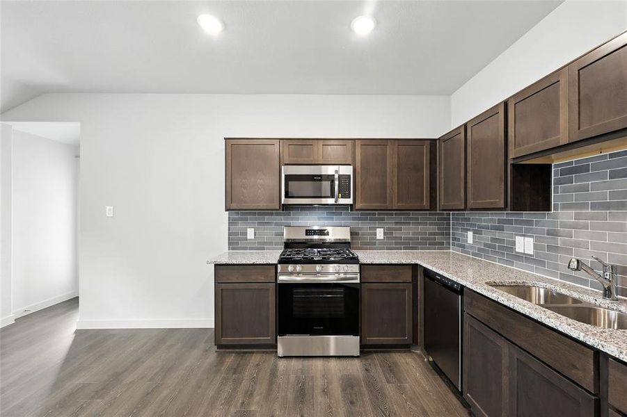 Kitchen with appliances with stainless steel finishes, dark brown cabinets, dark wood-style floors, tasteful backsplash, and light stone countertops Kitchen with appliances with stainless steel finishes, dark brown cabinets, dark wood-style floors, tasteful backsplash, and light stone countertops