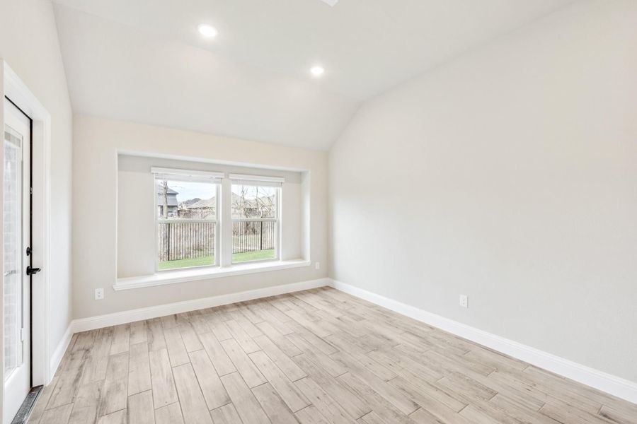 Empty room with vaulted ceiling, light wood floors, and large window overlooking backyard fence