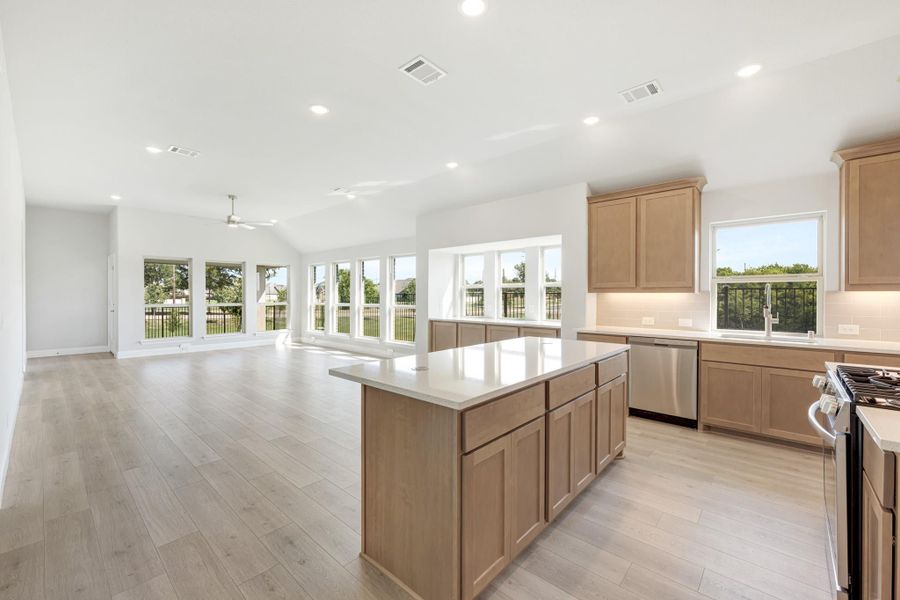 Open kitchen with light wood cabinets, center island, and stainless appliances flowing into a bright living area