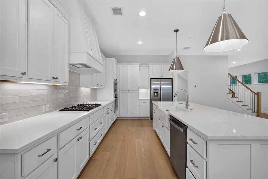 Modern kitchen featuring white shaker cabinetry, light wood-finish flooring, a central island with an integrated sink, stainless steel appliances, and pendant lighting Modern kitchen featuring white shaker cabinetry, light wood-finish flooring, a central island with an integrated sink, stainless steel appliances, and pendant lighting