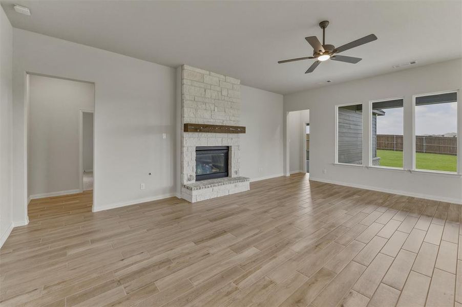 Spacious living area featuring a stone-clad fireplace with a wood mantel, wood-finish tile flooring, a ceiling fan, and multiple windows overlooking a private backyard with a wooden fence Spacious living area featuring a stone-clad fireplace with a wood mantel, wood-finish tile flooring, a ceiling fan, and multiple windows overlooking a private backyard with a wooden fence