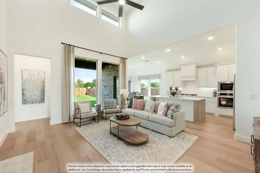 Open-concept living room with two-story ceiling, ceiling fan, hardwood floors, and kitchen visible in background.