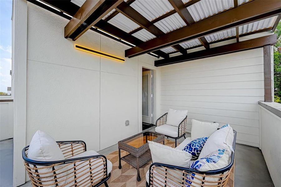 This cozy outdoor patio features stylish wicker seating with cushions and a glass-top coffee table. It's covered with a modern pergola, offering a charming space for relaxation or entertaining.