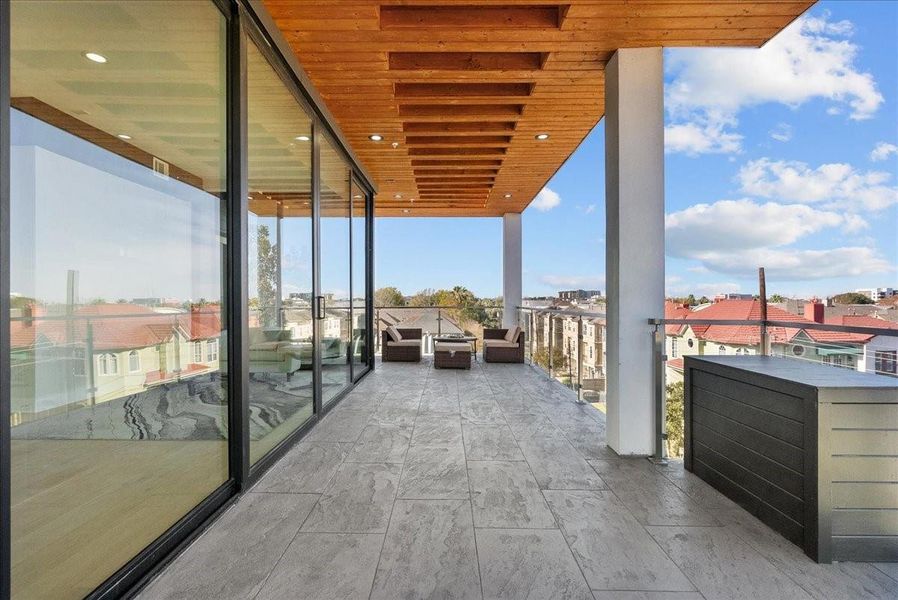 This photo showcases a spacious, modern balcony with sleek stone flooring and a wooden ceiling. It features comfortable outdoor seating, glass railings, and large sliding glass doors for seamless indoor-outdoor living, offering panoramic views of the surrounding neighborhood. This photo showcases a spacious, modern balcony with sleek stone flooring and a wooden ceiling. It features comfortable outdoor seating, glass railings, and large sliding glass doors for seamless indoor-outdoor living, offering panoramic views of the surrounding neighborhood.