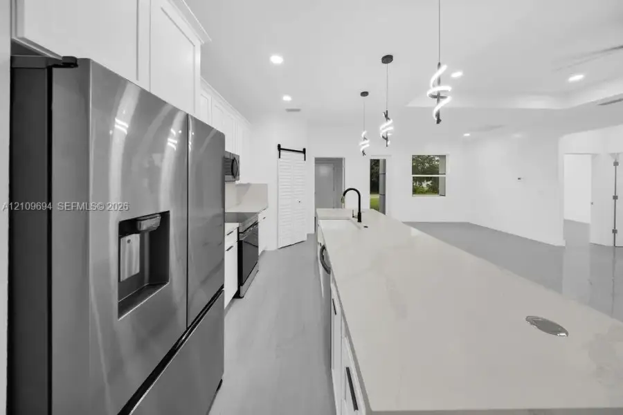 Kitchen featuring a spacious island with a neutral stone-surface countertop, white shaker-style cabinetry, stainless steel appliances, matte black hardware, and contemporary spiral pendant lighting