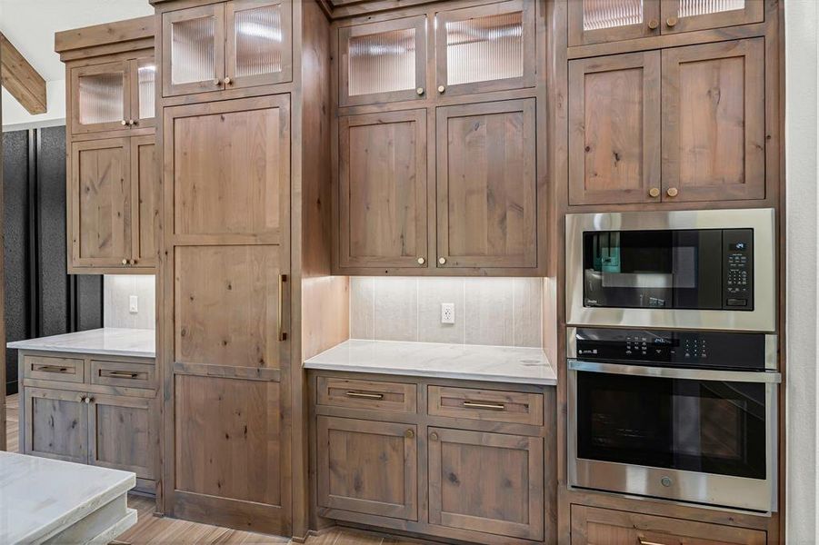 Kitchen with glass insert cabinets, stainless steel appliances, light stone counters, and brown cabinets Kitchen with glass insert cabinets, stainless steel appliances, light stone counters, and brown cabinets