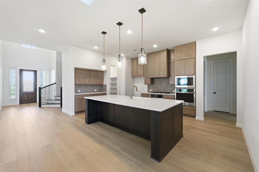 Open-concept kitchen featuring an oversized island with a white solid surface countertop, stainless steel appliances, light wood-finish cabinetry, and wood-finish flooring throughout