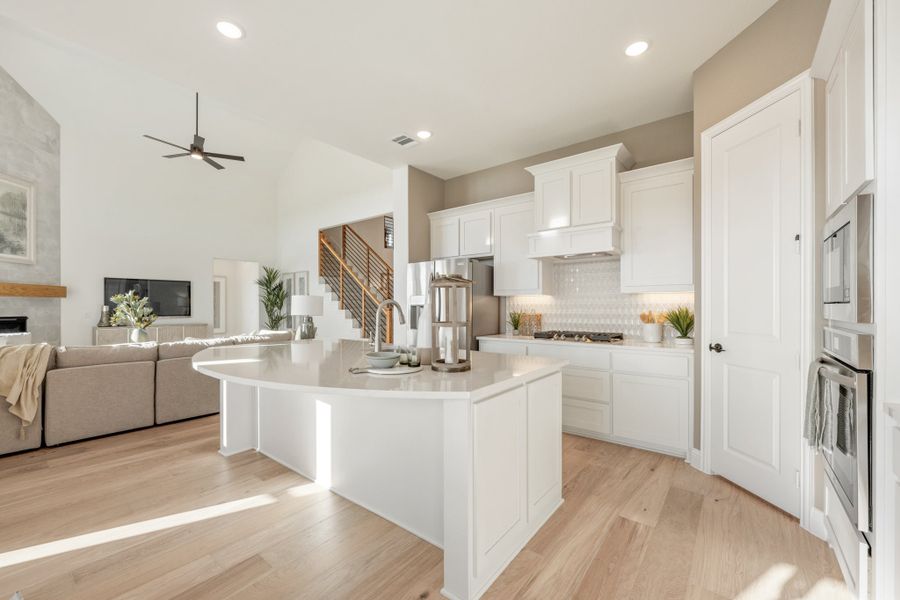 Open-concept kitchen with curved white island, white cabinets, light wood floors, and living room visible in background.