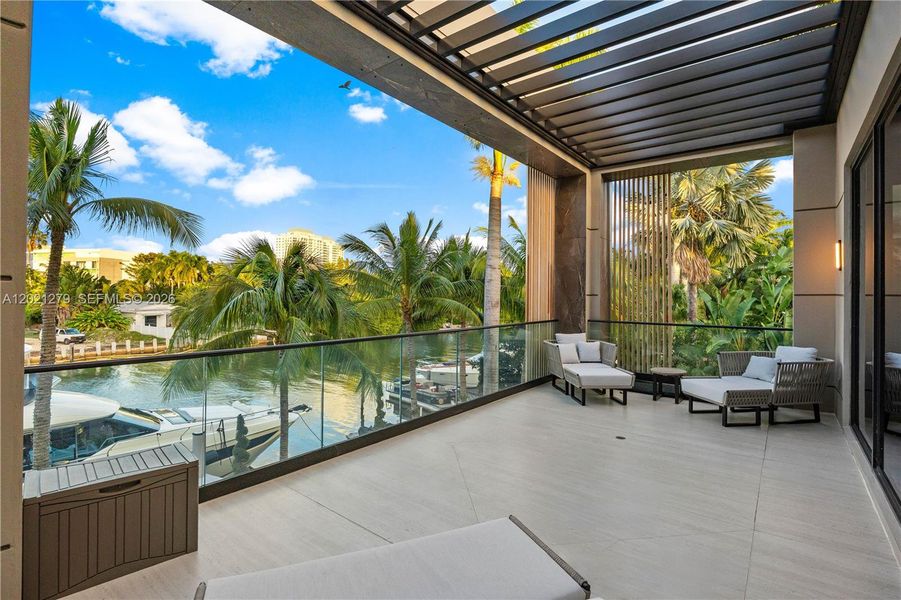 Second-floor waterfront balcony with motorized pergola, frameless glass railing, woven lounge chairs, large-format tile flooring, vertical wood slat screen, and unobstructed canal views with private dock and yacht below.