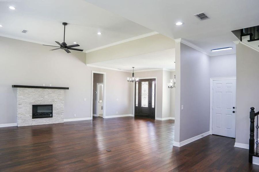 Unfurnished living room featuring a stone fireplace, ornamental molding, dark wood-style floors, ceiling fan, and a chandelier Unfurnished living room featuring a stone fireplace, ornamental molding, dark wood-style floors, ceiling fan, and a chandelier