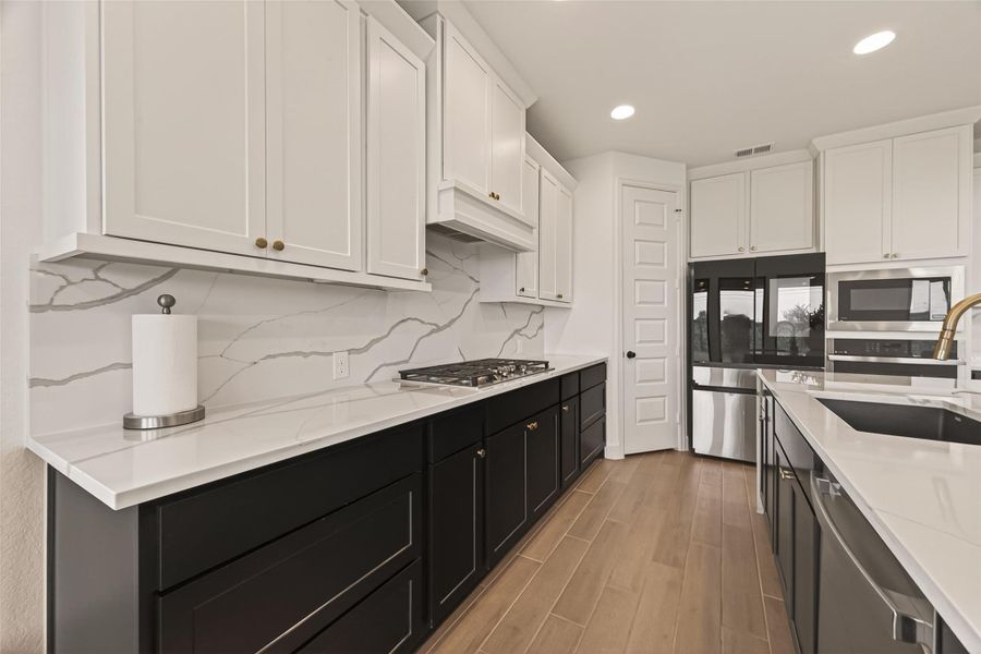 This modern kitchen features sleek Tricon black and Pearl white cabinetry, a marble-style backsplash, and stainless steel appliances. The room is well-lit with recessed lighting, and the design includes a spacious layout with ample countertop space.