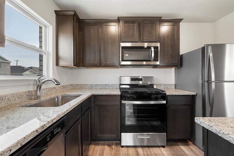 Kitchen with dark brown cabinets, light wood-type flooring, light stone counters, sink, and appliances with stainless steel finishes