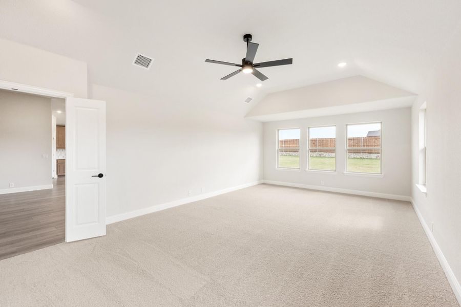 Empty bedroom with vaulted ceiling, ceiling fan, carpet flooring, and three windows overlooking backyard