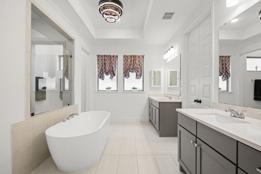 This spacious bathroom features a sleek freestanding tub, a large glass-enclosed shower, dual vanities with ample storage, and modern lighting. Natural light streams through two frosted windows, adding to the bright and airy feel.
