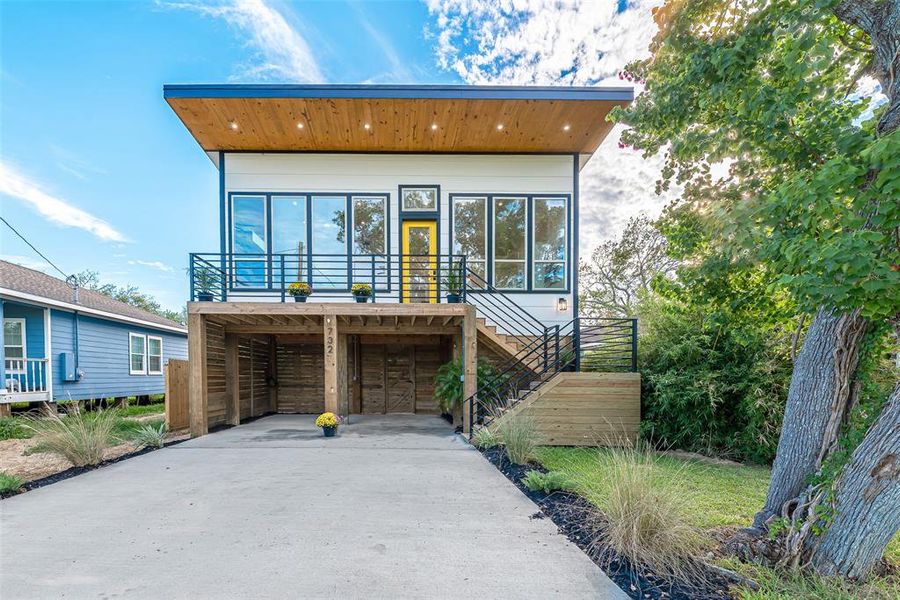 Modern two-story home with a strong geometric design, featuring a warm wooden upper exterior and a contrasting white lower half. The home includes a spacious raised deck with a metal railing, large windows for natural light, and covered parking space underneath.