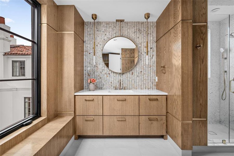 This ensuite bathroom is a true masterpiece, adorned with handsome wood paneling, elegant gold trim, a stunning stone backsplash, and a glass-encased, tiled walk-in shower, offering a blend of luxury and sophistication.