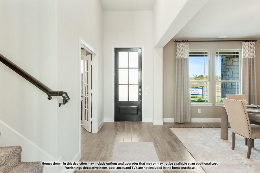 Bright home entryway with black front door, wood-look floors, staircase, and adjacent dining area with large windows.