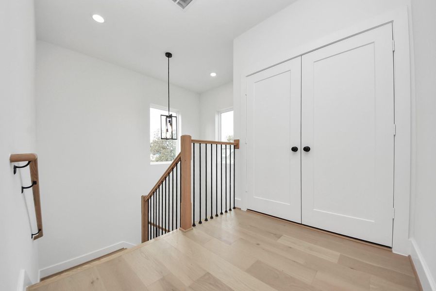 A bright landing area featuring light wood floors, white walls, and a sleek black metal railing. Pendant lighting, double closet doors, and large windows enhance the open, airy feel.
