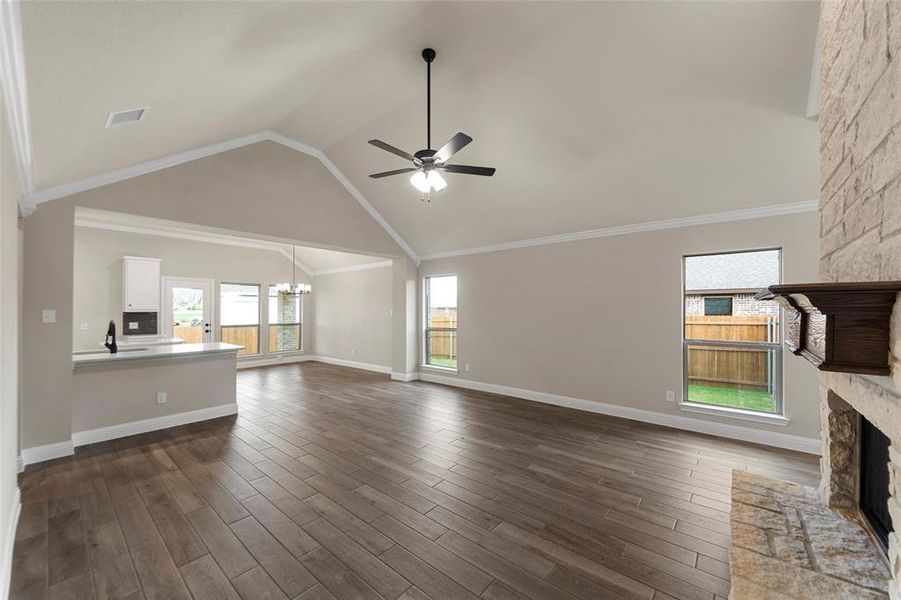 Expansive living area featuring vaulted ceilings, wood-finish flooring, a stone fireplace with a decorative wood mantel, multiple windows, and an open layout to the kitchen with a breakfast bar