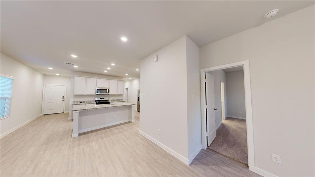 This photo showcases a modern, open-concept living space with light wood flooring and recessed lighting. The kitchen features white cabinetry, granite countertops, and stainless steel appliances. Adjacent is the primary bedroom.