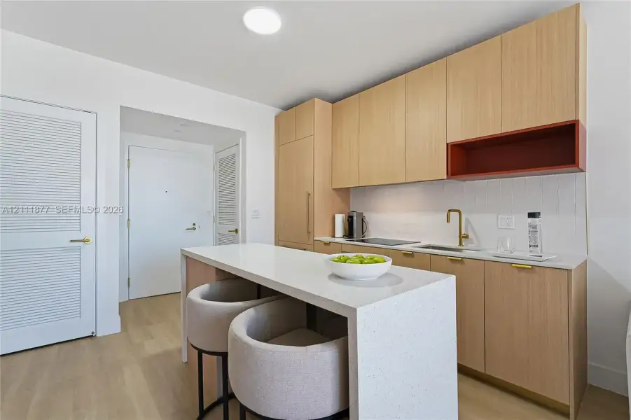 Kitchen featuring light-tone wood cabinetry, a solid-surface waterfall edge island, a contrasting open shelving unit, a brass-finish faucet, and white textured backsplash tile